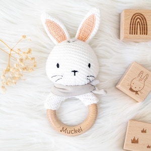 May include: White crocheted bunny rattle with a wooden ring and the name "Muckel" engraved on it. The rattle is surrounded by wooden blocks with different designs, including a rainbow, a bunny, and a crown.