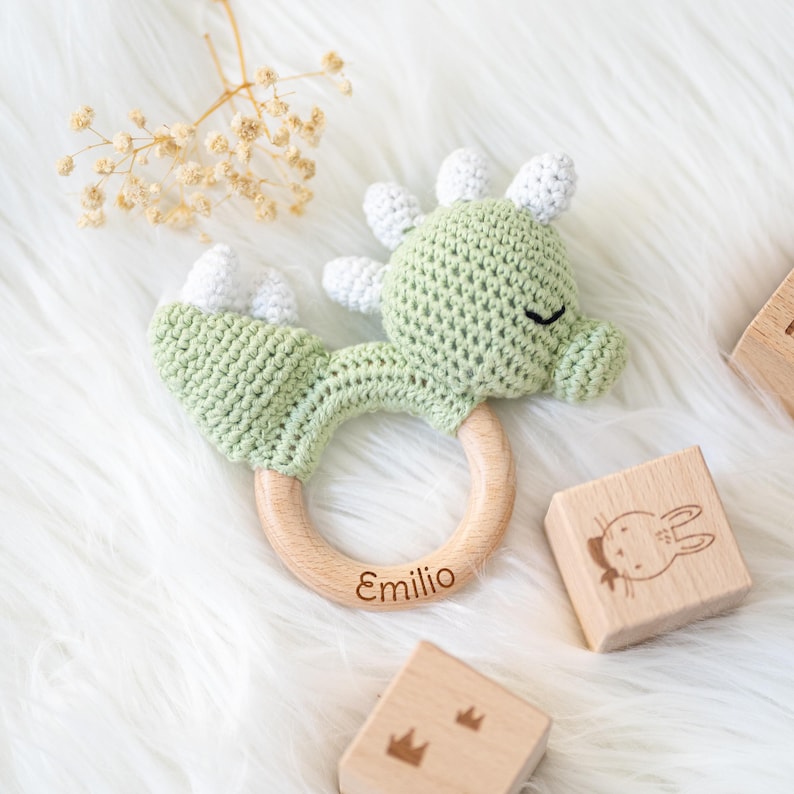 May include: A green crocheted dinosaur rattle with a wooden ring. The ring is engraved with the name "Emilio".