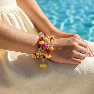 Könnte beinhalten: Ein mehrreihiges Armband mit gelben, rosa und transparenten Perlen. Ein großer gelber Anhänger hängt an dem Armband. Das Armband wird am Handgelenk einer Person getragen, mit einem cremefarbenen Kleid und einem blauen Wasserhintergrund.