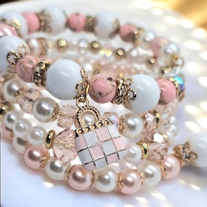 Könnte beinhalten: Mehrere Armbänder mit weißen, rosa und goldenen Perlen. Die Armbänder bestehen aus Perlen, runden Perlen und einem Handtaschen-Anhänger. Die Armbänder sind gestapelt, wodurch ein geschichteter Effekt entsteht. Die Perlen werden in Zentimetern gemessen.