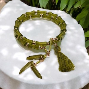 May include: A green bamboo-style bracelet with gold-colored accents and a tassel. The bracelet features a series of green, cylindrical beads connected by gold-colored links. A matching tassel and dangling green beads complete the design.