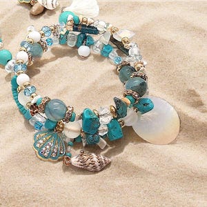Könnte beinhalten: Ein Armband mit Strandmotiv. Es hat türkisfarbene, weiße und transparente Perlen sowie Muschelanhänger. Das Armband ist mit einem großen weißen Muschelanhänger und kleinen Muschelanhängern verziert.