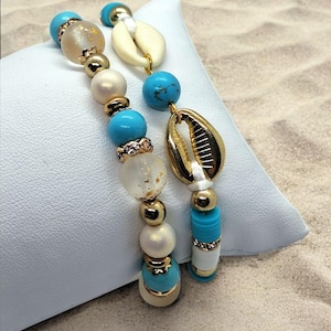 Könnte beinhalten: Zwei Armbänder mit Perlen im Strand-Thema. Ein Armband hat türkisfarbene, goldene und perlenartige Perlen sowie einen goldfarbenen Muschelanhänger. Das andere Armband hat ähnliche Perlen und einen weißen Muschelanhänger. Die Armbänder werden auf einem weißen Kissen präsentiert.