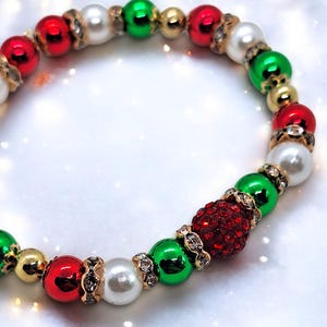Könnte beinhalten: Ein festliches Armband mit roten, grünen, weißen und goldenen Perlen. Das Armband kombiniert glänzende und perlmuttartige Perlen mit einer zentralen roten, strassbesetzten Perle. Goldfarbene Abstandshalter mit klaren Strasssteinen sorgen für Glanz.
