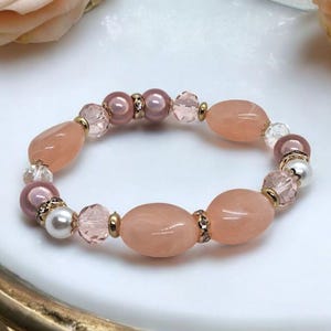 Könnte beinhalten: Ein Armband mit pfirsichfarbenen, ovalen Perlen, ergänzt durch kleinere, runde und kristallartige Perlen. Goldfarbene Abstandshalter und weiße Perlen setzen Akzente. Das Armband ist zart und feminin gestaltet.