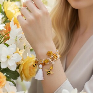 May include: A multi-strand bracelet with yellow, gold, and white beads. The bracelet features bee and honeycomb charms, and a white tassel. The bracelet is worn on a wrist, with a floral background of yellow and white flowers.