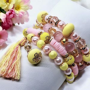 Könnte beinhalten: Mehrere Armbänder mit gelben, rosa und Perlenperlen. Die Armbänder haben goldfarbene Akzente und eine Quaste mit gelben und rosa Fäden. Ein Strauß rosa und gelber Blumen befindet sich im Hintergrund.
