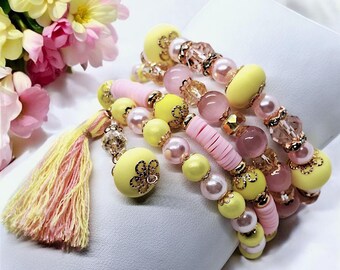 May include: A collection of beaded bracelets in shades of yellow, pink, and pearl. The bracelets feature various bead shapes, including round, cylindrical, and faceted, with gold-colored accents. A matching tassel with yellow and pink threads is also visible.
