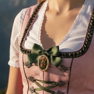 Könnte beinhalten: Ein rosafarbenes Dirndl mit einer weißen Bluse mit Spitzenbesatz am Ausschnitt, einem dunkelgrünen Mieder mit Band-Schnürung und dekorativen Metallösen sowie einer ovalen Brosche mit kleiner Schleife und Perlenrand an der Brust.