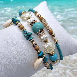 Könnte beinhalten: Drei Armbänder mit Perlen im Strand-Thema. Die Armbänder haben türkisfarbene, weiße und goldfarbene Perlen sowie Muscheln. Die Armbänder werden auf einem weißen Kissen mit verschwommenem Meereshintergrund präsentiert.