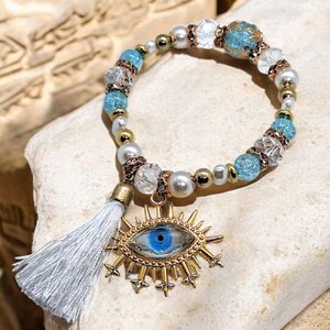 May include: A beaded bracelet with a gold evil eye charm, blue iris, and white tassel. The bracelet has blue, white, and clear beads, plus gold and pearl accents. This bracelet is a fashion accessory.