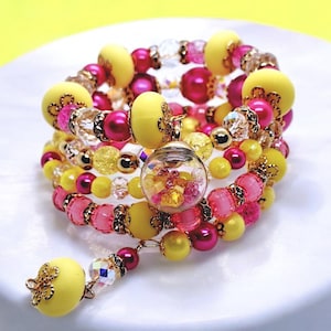 Könnte beinhalten: Ein mehrsträngiges Armband mit gelben, rosa und transparenten Perlen. Das Armband enthält einen Anhänger in Form einer durchsichtigen Kugel, gefüllt mit bunten Akzenten, und eine baumelnde gelbe Perle mit goldfarbenen Akzenten. Die Perlen sind spiralförmig angeordnet.