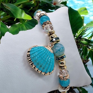 Könnte beinhalten: Ein Armband mit einer türkisfarbenen Muschelanhänger. Das Armband hat goldfarbene Perlen, klare und blaue Kristallakzente sowie hellblaue und weiße Perlen. Der Muschelanhänger hat eine goldfarbene Umrandung.