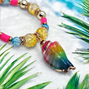 Könnte beinhalten: Ein farbenfrohes Armband mit einem Muschel-Anhänger. Das Armband hat rosa, gelbe, blaue und goldene Perlen. Der Muschel-Anhänger ist mit einem Farbverlauf aus Rosa, Gelb, Grün und Blau bemalt und hat goldene Verzierungen.