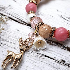 Puede incluir: Una pulsera de cuentas rosas, blancas y de madera, acentuada con espaciadores dorados. Tiene un dije de ciervo dorado y un dije de flor blanca. La pulsera está sobre una superficie de madera blanca.