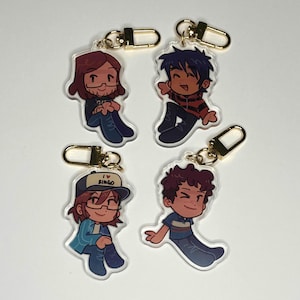 Peut inclure: Quatre porte-clés en acrylique avec des illustrations de personnages de dessins animés. Chaque porte-clés a un fermoir doré. Les personnages sont représentés dans diverses tenues et poses, avec des détails comme des lunettes et des chapeaux. Un porte-clés porte l'inscription "I ❤️ BINGO".