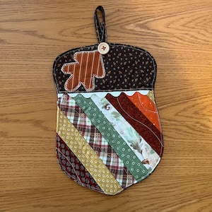 May include: A quilted fabric potholder shaped like an acorn with a dark brown top featuring a small orange leaf applique and a light-colored button. The bottom section consists of diagonal fabric strips in shades of green, brown, and orange.