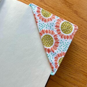 May include: A triangular fabric corner bookmark featuring a white background with a repeating pattern of orange-petaled sunflowers with olive green centers, accented by small teal polka dots, fitted over the corner of a white page.