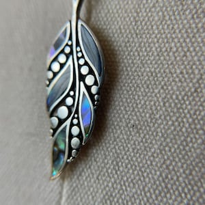 Può includere: Un ciondolo d'argento a forma di foglia con intarsi di madreperla di abalone iridescente. La foglia ha un design dettagliato con piccoli cerchi e linee.