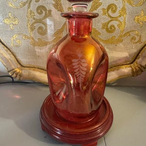 Puede incluir: Decantador de vidrio rojo rubí con tapón a juego, sobre una base de madera. El decantador presenta un diseño de hojas y probablemente se utiliza para licores o decoración.