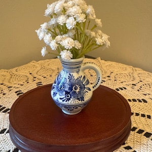 May include: A small ceramic pitcher featuring a white base with blue floral patterns and a single handle, filled with a bouquet of small white artificial flowers. The pitcher has a rounded body and a flared rim, decorated with hand-painted blue petals.