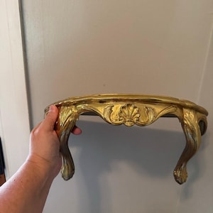 May include: A gold-colored wall shelf featuring a curved, semi-circular top and two ornate, carved legs. The front apron of the shelf is decorated with a central shell motif flanked by scrolling leaf patterns, finished with a metallic gold paint.