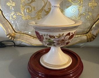 May include: A white ceramic lidded serving dish with floral designs in shades of purple and brown, accented with gold trim. The dish sits on a dark red wooden stand. The lid is topped with a gold finial.