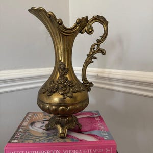 May include: A gold-colored decorative pitcher with ornate floral and scrollwork details. The pitcher has a flared spout and a curved handle. It is displayed on a stack of books, including "Reese Witherspoon Whiskey Teacup" and "Southern Living Party Cookbook."