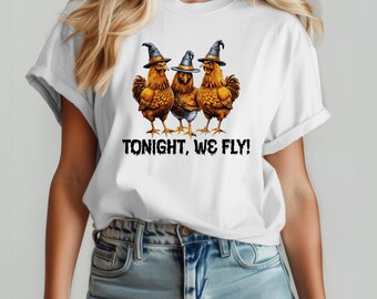 May include: A white t-shirt featuring a graphic of three brown chickens wearing black witch hats. Below the chickens, the text reads TONIGHT, WE FLY! in a black, distressed, spooky font. The shirt has short sleeves and a crew neckline.