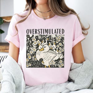 K&ouml;nnte beinhalten: Hellrosa T-Shirt mit dem Wort "OVERSTIMULATED" &uuml;ber einer Grafik einer Ente, umgeben von Entenk&uuml;ken und Blumen. Die Grafik ist in Schwarz, Wei&szlig; und Gelb gehalten.