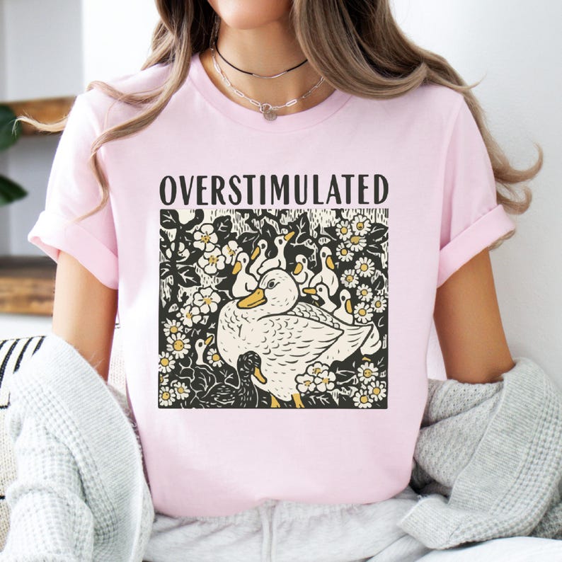 K&ouml;nnte beinhalten: Hellrosa T-Shirt mit dem Wort "OVERSTIMULATED" &uuml;ber einer Grafik einer Ente, umgeben von Entenk&uuml;ken und Blumen. Die Grafik ist in Schwarz, Wei&szlig; und Gelb gehalten.