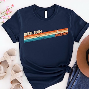 May include: Navy blue t-shirt with the text "REBEL SCUM" above a retro-style design with the text "SINCE 1977". The design features horizontal stripes in orange, yellow, and teal. The shirt is knotted at the bottom.