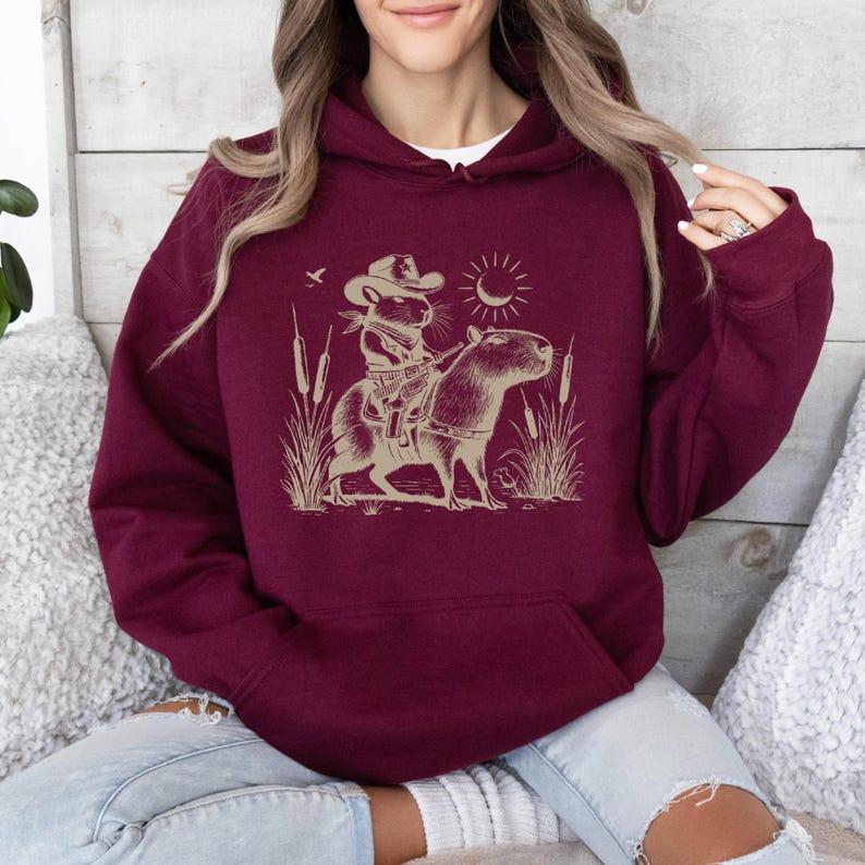 K&ouml;nnte beinhalten: Ein maroonfarbener Hoodie mit einem Aufdruck eines Cartoon-Nagetiers, das einen Cowboyhut tr&auml;gt und ein Capybara reitet. Der Aufdruck ist im Vintage-Stil mit einer Sonne und V&ouml;geln im Hintergrund.