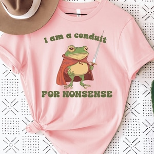 May include: Pink t-shirt featuring a cartoon frog in a red cape holding a sword. The text "I am a conduit FOR NONSENSE" is printed above and below the frog illustration. The shirt has short sleeves and a crew neck.