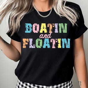 Puede incluir: Camiseta negra con las palabras "BOATIN and FLOATIN" en letras coloridas y desgastadas. El texto est&aacute; en un arco&iacute;ris de colores pastel, incluyendo azul, rosa, amarillo, naranja y morado. La camiseta tiene mangas cortas.