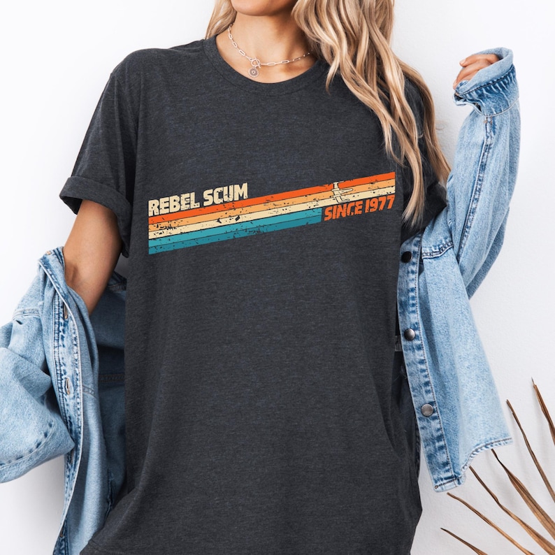 May include: Dark gray t-shirt with the text "REBEL SCUM" and "SINCE 1977" in a retro-style design. The design features horizontal stripes in orange, yellow, blue, and teal. A casual, vintage-inspired graphic tee.