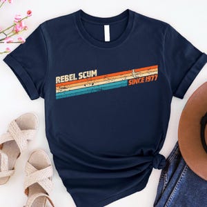 May include: Navy blue t-shirt with the text "REBEL SCUM" above a retro-style design with the text "SINCE 1977". The design features horizontal stripes in orange, yellow, and blue.