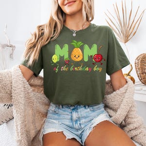 May include: Olive green t-shirt with the word "MOM" in green letters and a pineapple in the center. The shirt features cartoon fruit characters and the text "of the birthday boy" in colorful embroidery.