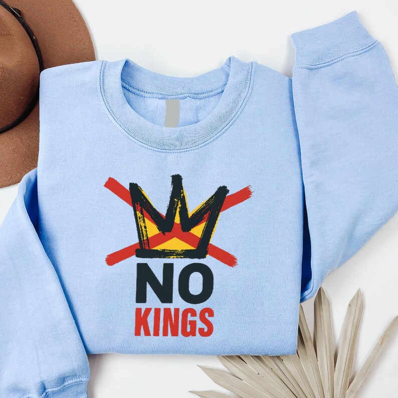 May include: Light blue sweatshirt with a graphic of a crown crossed out with red brushstrokes. Below the crown, the words "NO KINGS" are printed in black and red. The sweatshirt is made of a soft material.