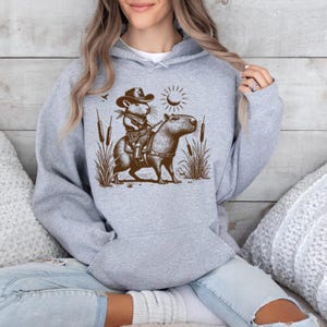 K&ouml;nnte beinhalten: Ein grauer Kapuzenpullover mit einer braunen Illustration einer Maus, die einen Cowboyhut tr&auml;gt und ein Capybara reitet. Die Illustration ist vor einem Hintergrund aus Schilf und einer Sonne.