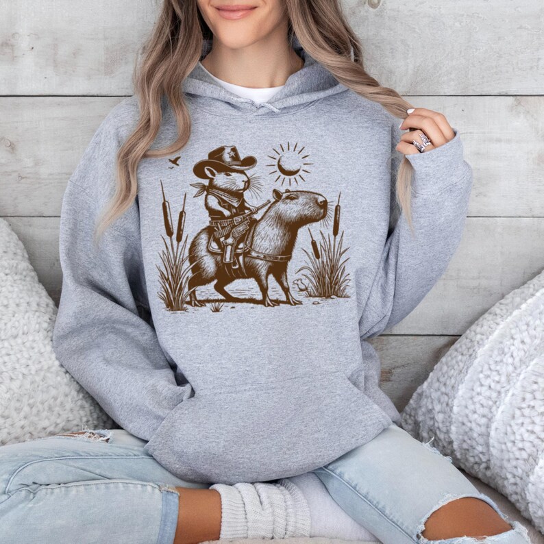 K&ouml;nnte beinhalten: Ein grauer Kapuzenpullover mit einer braunen Illustration einer Maus, die einen Cowboyhut tr&auml;gt und ein Capybara reitet. Die Illustration ist vor einem Hintergrund aus Schilf und einer Sonne.