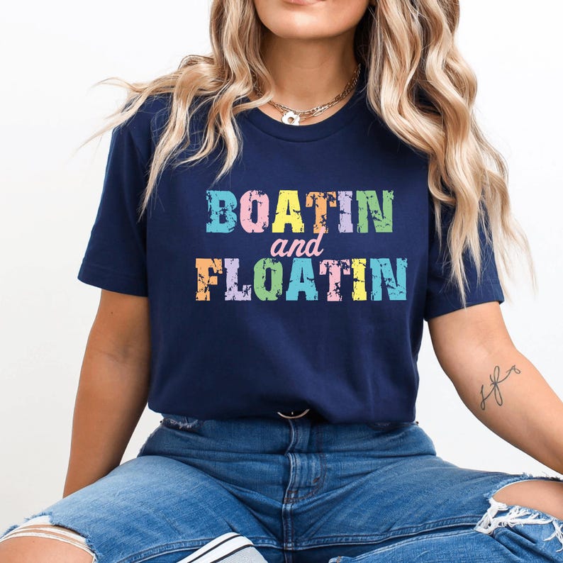 Puede incluir: Camiseta azul marino con las palabras "BOATIN and FLOATIN" en letras coloridas y texturizadas. La camiseta tiene cuello redondo y mangas cortas. El texto est&aacute; en una paleta de colores pastel.