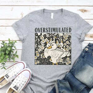 K&ouml;nnte beinhalten: Graues T-Shirt mit dem Wort "OVERSTIMULATED" &uuml;ber einer Grafik einer Ente mit Entenk&uuml;ken, umgeben von Blumen. Wei&szlig;e Leinenschuhe und blaue Jeans sind ebenfalls zu sehen.