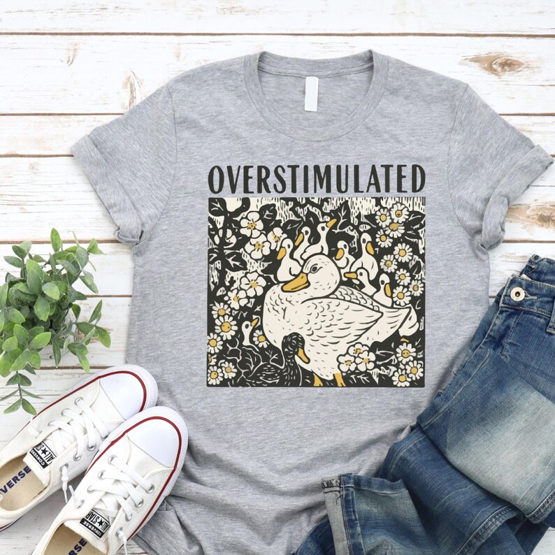 K&ouml;nnte beinhalten: Graues T-Shirt mit dem Wort "OVERSTIMULATED" &uuml;ber einer Grafik einer Ente mit Entenk&uuml;ken, umgeben von Blumen. Wei&szlig;e Leinenschuhe und blaue Jeans sind ebenfalls zu sehen.