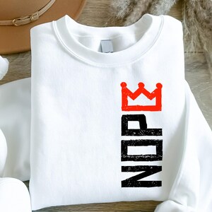 May include: White sweatshirt with a red crown graphic above the black letters "NDN". The sweatshirt is a crew neck style and appears to be made of a soft material. The design is printed on the front of the shirt.