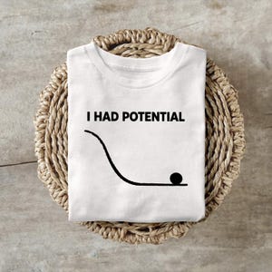 May include: White t-shirt with the text "I HAD POTENTIAL" above a black line drawing of a ball at the bottom of a curved ramp. The shirt is folded and resting on a woven, circular basket.