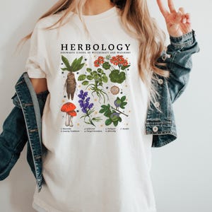 May include: Cream-colored t-shirt featuring a botanical illustration with the text "HERBOLOGY HOGWARTS SCHOOL OF WITCHCRAFT AND WIZARDRY." The design includes drawings of various plants, including a mandrake, mushrooms, and flowers.