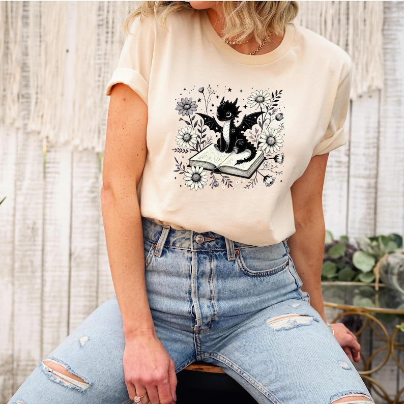 May include: A beige t-shirt featuring a black and white graphic of a dragon sitting on an open book, surrounded by flowers. The dragon has large wings and is facing forward. The t-shirt is paired with blue jeans.