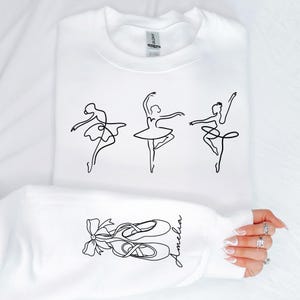 May include: White sweatshirt featuring three line-drawn ballerinas in various poses. The sleeve displays a line drawing of ballet shoes with a bow and the name "Amelia" in script. The sweatshirt is made of a soft material.