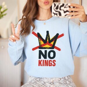May include: A light blue sweatshirt with a graphic design featuring a crown crossed out with red lines, and the words "NO KINGS" in black and red. The sweatshirt is a casual style.